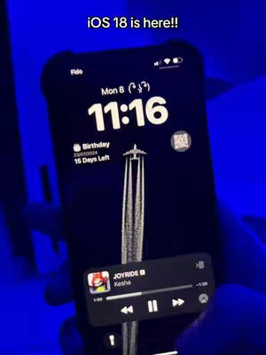 A person holding a black iPhone displaying the home screen with a custom wallpaper and a music player interface. The screen is in focus, set against a blurred blue backdrop, likely indoors. The phone shows the time, date, and widgets, indicating a focus on iOS customization.