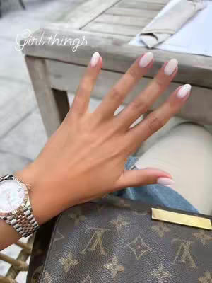 A close-up shot of a hand with well-manicured nails, a watch, and a designer handbag. The hand is in focus, with light pink nail polish. The watch has a silver and gold band. The handbag is brown with the iconic LV monogram. The background is slightly blurred, suggesting a casual setting, with text overlay: Girl things.