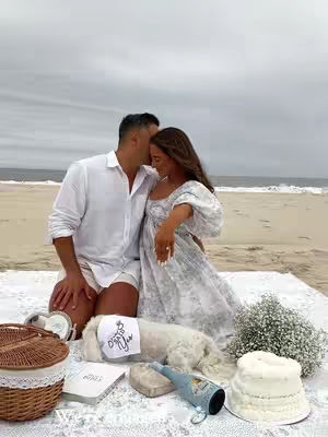 A couple is posing on a beach. The man is wearing a white button-down shirt and white shorts. The woman is wearing a white floral dress and has an engagement ring on her finger. They are on a white blanket with a picnic basket, flowers, and a cake. A small dog is also present wearing a headband.