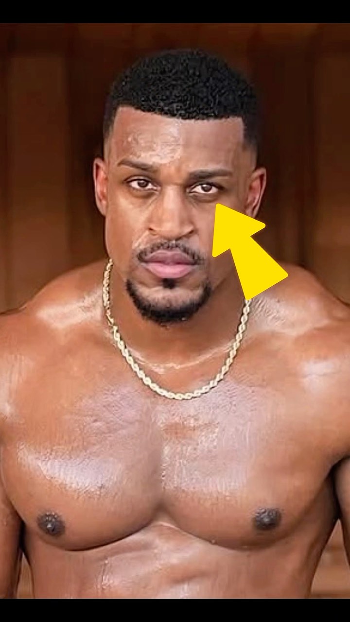 A shirtless man with a muscular physique and a gold chain gazes intensely at the viewer. A yellow arrow points at his eye. The background is a blurred wooden texture.