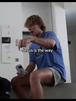 A young man with short, light brown hair is sitting and addressing the camera. He is wearing a blue t-shirt and gray shorts. Text overlayed on the image reads, "Jesus is the way."
