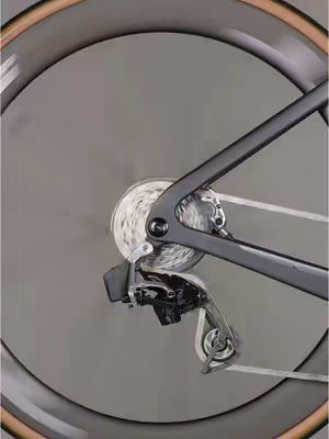 The image shows the rear wheel of a bicycle. The wheel is in motion, with the spokes blurred due to the speed of rotation. The bicycle's frame and the rear derailleur are visible. The background is a uniform gray, which emphasizes the bike parts.