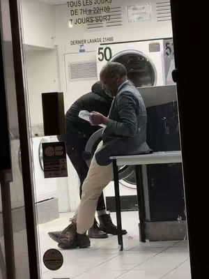 A man in a grey jacket and khaki pants sits at a table inside a laundromat, reading a paper. Another person in black clothing stands nearby. The laundromat has white washing machines and a glass door with French text.