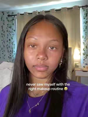 A woman with long dark hair in a purple shirt looks directly at the camera. The background includes curtains and a wall with a leafy design. Text overlayed on the image reads, 'never saw myself with the right makeup routine'.
