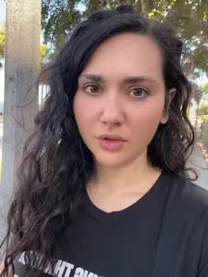 A woman with dark hair and a black t-shirt stands outside, likely in a park or urban setting; the background is blurred, with trees and a sidewalk visible. The focus is on her face, which appears to be close up. Light appears natural.
