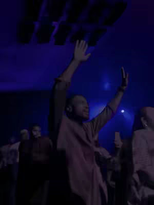 A dark, vertical image captures a live performance with a man in the foreground raising his arms in worship. The scene is dimly lit with a predominant blue and purple color scheme. Behind him, other figures are visible, also appearing to be engaged in worship.