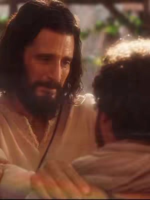 The image shows a scene with two men. The main focus is on one man, presumably Jesus, who has long dark hair and a beard and is wearing a cream-colored robe. He is looking down with a gentle expression. The background is blurred and suggests a natural outdoor setting. The second man is cropped, but appears to be listening and receptive to the first figure.
