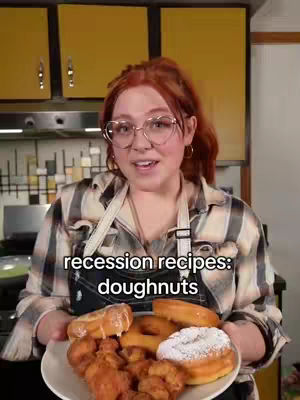 A woman with red hair and glasses is holding a plate of doughnuts in a kitchen setting. She is wearing an apron and flannel shirt. The text overlay indicates the video is about 'recession recipes' and focuses on doughnuts.