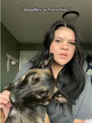 A woman with dark hair and a nose piercing is holding a dog close to her face. The dog appears to be a mix with tan, brown and black markings. The woman is wearing a grey t-shirt in a room with olive green walls and a white fixture.