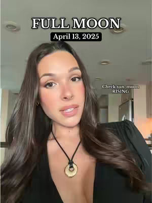 A woman with long brown hair and a black top is centered in the frame, with text overlays at the top reading 'FULL MOON' and 'April 13, 2025'. The background is blurred but suggests an interior setting, maybe a kitchen with soft warm lighting and a view of cabinets and a refrigerator. The woman is looking at the camera and speaking.