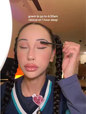 A close-up shot of a woman applying mascara while looking in a mirror. She has braided pigtails, full lips with visible lip gloss, and is wearing a navy blue top with white and teal accents. The background is softly lit with warm tones. Text overlayed on the image reads: 'grwm to go to 6:30am clinical on 1 hour sleep'. A heart-shaped charm is visible on her chest.
