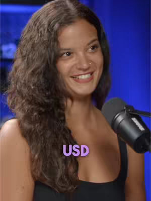 A smiling woman with wavy brown hair is centered in the frame. She has a nose ring and is wearing a black top. Partially visible is a black microphone to her right, against a blurred blue background. The word 'USD' appears in a purple box near her chest.