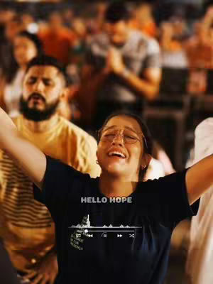 A young woman with glasses and arms outstretched, appearing to be in worship. She has her eyes closed and mouth open, showing an expression of joy or reverence. She's standing among a crowd of people, some with their hands raised as well, in what appears to be a church or religious gathering. The background has warm lighting, adding to the emotional atmosphere.
