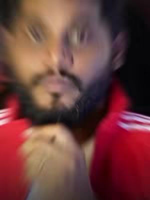 The image shows a close-up of a man's face with a blurred effect. The man has a dark beard and is wearing a red jacket with white stripes on the shoulders. The background is dark and out of focus, enhancing the focus on the man's face.