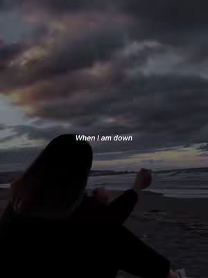 A silhouette of a person is standing on a beach with their back to the camera, looking out at the ocean. The sky is filled with dark, moody clouds. Text overlayed on the image reads, 'When I am down'.