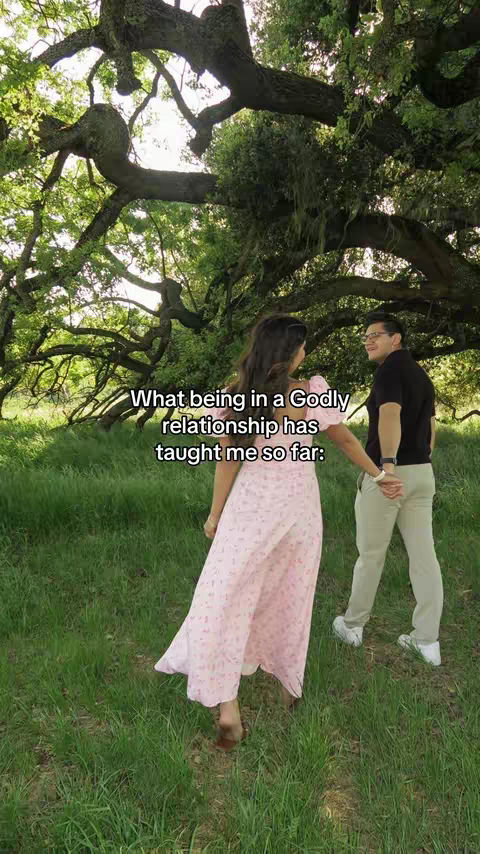 A couple walks hand-in-hand through a sunlit grassy area towards the viewer. The woman is barefoot, wearing a long pink floral dress. The man, wearing glasses, walks beside her, and is wearing a black shirt and beige pants. Text overlayed on the image reads, 'What being in a Godly relationship has taught me so far:'