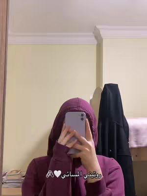 A person wearing a maroon hoodie is holding a phone in front of their face, partially obscuring it. The background is a lightly-colored wall with a white trim at the ceiling. A dark coat is hanging to the right, and a wooden border is visible on the left.