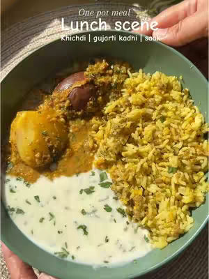 Close-up shot of a plate containing a portion of yellow rice, what appears to be a curry with a whole eggplant and a cut potato, and a white liquid with green herbs. The plate, which the food is on rests on a surface with a woven pattern. The lighting appears to be natural, with soft shadows.
