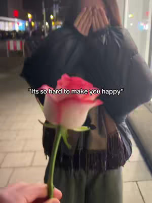 A hand holding a pink and white rose is shown in the foreground. Behind, a person is seen from the back, wearing a black jacket, with their hands up covering their face. In the background, there are buildings and street lights, suggesting a night scene.