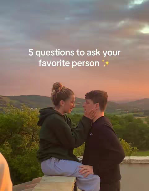 A young woman and a young man face each other intimately against a backdrop of a beautiful sunset. The woman is holding the man's face tenderly while they look deep in each other's eyes. The sky is a gradient of pinks and oranges.
