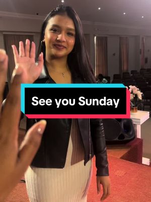 A person with long, dark hair waves at the camera with their right hand. They are wearing a black leather jacket over a cream-colored dress and gold hoop earrings. In front of them is another person's hand, and the background shows a church interior with rows of chairs and a stage. A blue and red rectangular text box reads "See you Sunday."