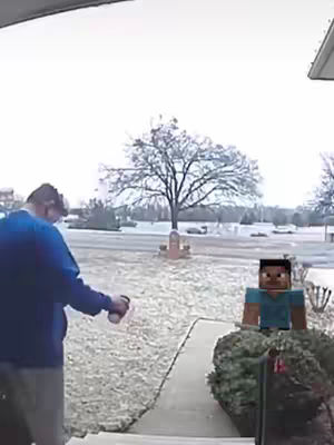 A person in a blue shirt is facing away from the camera, spraying something, likely water, onto a lawn covered in snow. To the person’s right, the Minecraft character “Steve” is digitally added to the image, standing in front of a small bush. The background shows a snowy landscape, a house, a bare tree, and a pale, overcast sky.