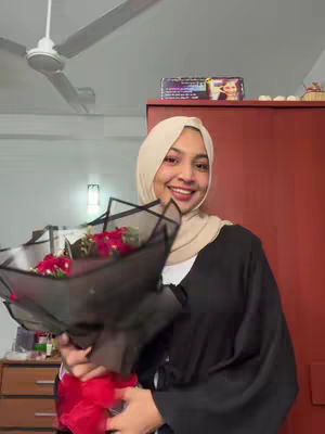 A smiling woman in a beige hijab and black cardigan holds a bouquet of red roses. She is indoors, with a ceiling fan visible above and furniture behind her. The lighting is natural.