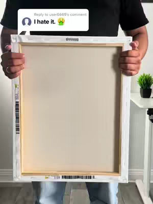 A person holding the back of a canvas, the canvas is white with the wooden frame and its back is exposed. Overlayed on the top-left is a comment box. Behind the person is a white desk with a small plant on it, providing a neutral background.