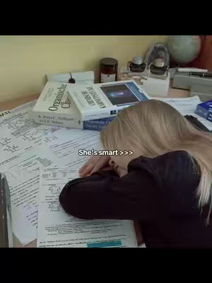 A woman is resting her head on a desk covered in papers and books. Overlaid text reads, 'She's smart >>>'. The scene suggests exhaustion related to academic work.