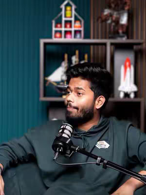 A man wearing a hoodie sits in front of a microphone, looking slightly to the side. Behind him is a teal wall and a shelf with decorative items.