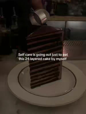 A vertical shot of a layered cake on a white plate, with a dark background. The cake is tall and rectangular with multiple layers, predominantly chocolate. The plate is plain white with a gold rim detail. The background is dark, possibly a restaurant or cafe setting, with some out-of-focus shapes.