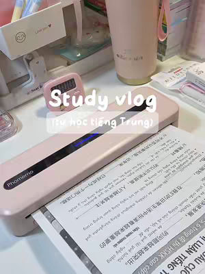 A pastel-colored desk setup with study materials. A pink laminator is in the foreground, with a sheet of paper with Chinese characters coming out of it. The text 'Study vlog (từ học tiếng Trung)' appears on top of a pink digital timer. Other items include a light pink tumbler, pens, a box with 'Love you' written on it.