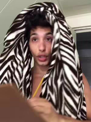 A young man is shown in a selfie-style video. He is looking at the camera with his face framed by a black and white zebra-striped towel wrapped around his head. A yellow pencil is seen held near his neck, and his bare chest is visible.