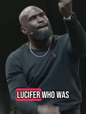 The image presents a bald, dark-skinned man with a beard, wearing a black sweater and a silver necklace. He's gesturing upwards with his right hand, facing slightly to his left. Overlay text in a red box presents the words "LUCIFER WHO WAS".