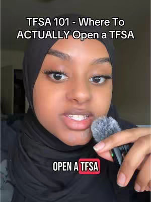 A close-up shot of a person with dark skin, wearing a black hijab. The person is looking directly at the camera, and in front of their face is a microphone. Text overlay at the top reads "TFSA 101 - Where To ACTUALLY Open a TFSA", and a second text overlay on the bottom reads "OPEN A TFSA". The background is blurred, showing a soft, neutral color and possible lighting.