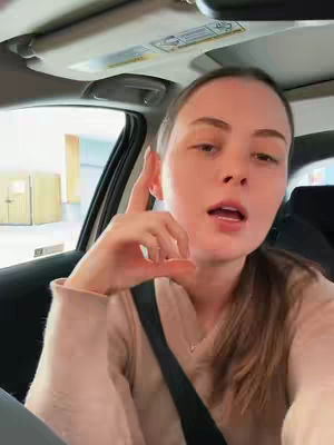 A woman is seated inside a car, angled slightly towards the camera. She has light skin, long brown hair pulled back and a light pink shirt. The background reveals a building. The interior of the car is visible with the seat belt and the car's ceiling light.