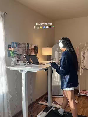 A young woman stands on a treadmill desk setup in her home office. She is wearing a dark blue sweater, white shorts, and white headphones. The room is bright and has neutral-colored walls. A standing desk with a laptop and tablet and a mirror behind her, indicating a bedroom setup.