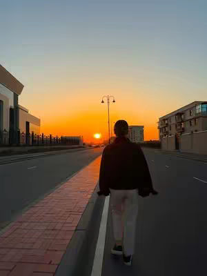 A person is walking away from the camera down a road with a sunset in the background. The sky is filled with orange and the sun is setting in the middle of the frame. There are buildings on either side of the road. The person is wearing a black jacket and white pants.
