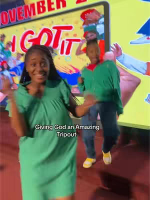 A woman in a green dress is the focal point, dancing and smiling. Behind her, a man in a green jacket dances, and a large screen displays text and images. The overall scene suggests a lively and energetic atmosphere, possibly a church performance or a similar event.