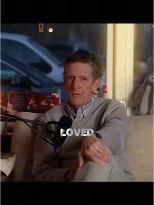 A man sits in an armchair, facing the camera. He wears a light-colored sweater over a button-down shirt. The background has a window with a blurry street view.