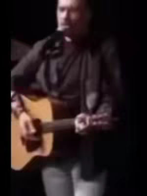 A blurry, low-quality image shows a man playing an acoustic guitar and singing into a microphone during a performance. The background is dark, focusing attention on the performer, who is wearing dark clothing and holding the guitar in a typical performance posture.