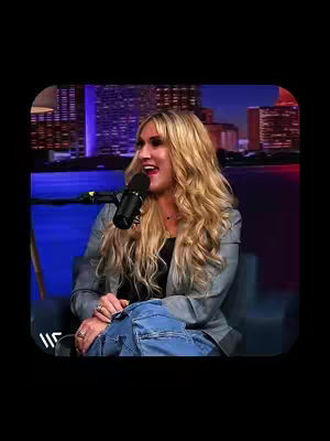 A woman with long blonde hair sits in a chair, speaking into a microphone. She is in a studio setting with a cityscape visible in the background. Overlayed text indicates the video's motivational and inspirational theme.