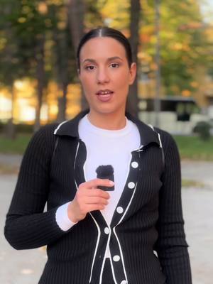 A woman stands outdoors against a blurred background, holding what appears to be a black microphone. She is wearing a white shirt under a black cardigan with white trim and buttons. The background is green, indicating trees or foliage.