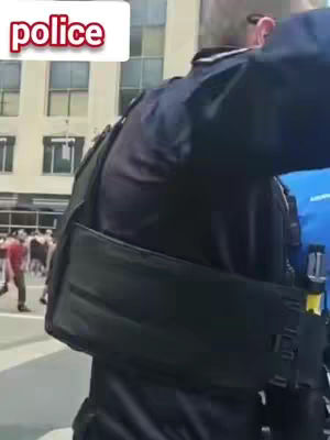 The image shows a medium shot taken from behind a police officer. The officer is wearing a dark blue uniform with a tactical vest. The word "police" is displayed in a white box with a red outline in the upper-left corner of the image. In the background, a slightly blurred street scene and a crowd of people adds to the ambiance.
