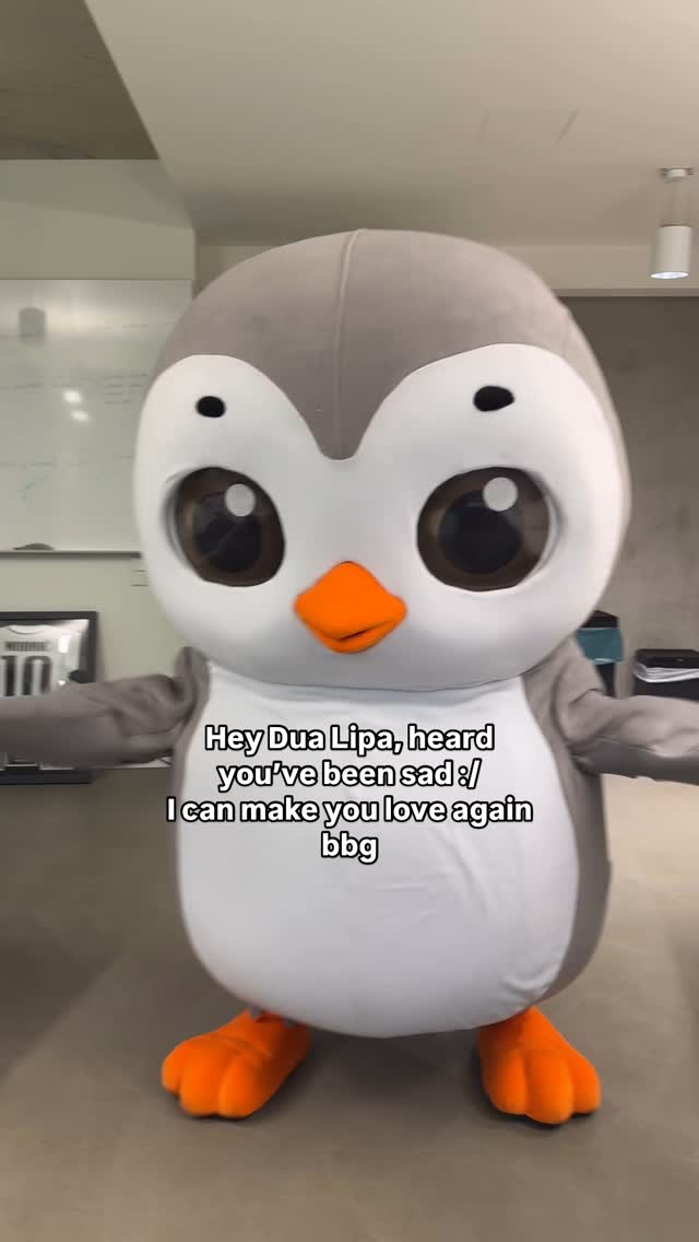 A person in a penguin costume stands in a room. The penguin is gray and white with an orange beak and feet. Text overlay on the image includes a message for Dua Lipa.