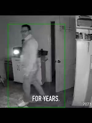 A security camera image in grayscale shows a man inside a room, framed by a green box, implying a recording. The man, wearing glasses and a light-colored shirt, is walking away from the camera, with his back partially turned. The background includes a cabinet, door, and kitchen items. In the bottom part of the image, text overlays: 'FOR YEARS.'