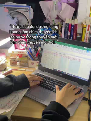 The image captures a workspace with a laptop open, displaying a document. The hands are typing on the keyboard. Other elements include a cluttered desk with papers, pens, and other stationary, as well as books, a fan and a clock.