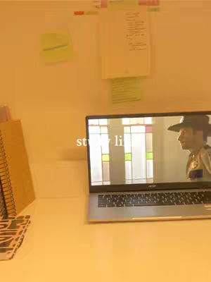 The image shows a laptop on a white desk.  The screen displays a scene with a person in a sheriff's hat and a colorful, stained-glass window effect.  On the left side of the frame, there are notebooks and other stationery items. The background is a warm, yellow-toned color, possibly from the lighting.