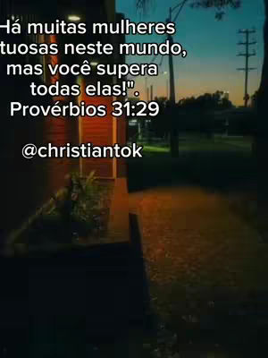 The image is a vertical shot with blurred background. The foreground features a pathway with a blurry building on the left and a dark tree and streetlights on the right. Overlayed on the image is white text in Portuguese, a bible verse and the handle of the account.