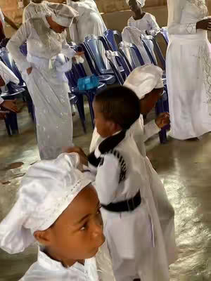 Several people in white robes and head coverings are gathered inside, looking at each other. The image is slightly low-angle, capturing the group from a medium distance, with some individuals in the foreground and others in the background. The lighting appears consistent throughout the interior.